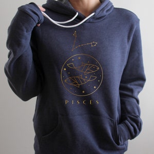 May include: A navy blue hoodie with a gold outline of two fish and stars in a circle. The word "PISCES" is printed below the circle.