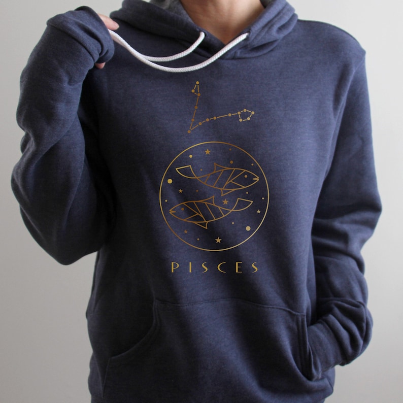 May include: A navy blue hoodie with a gold outline of two fish and stars in a circle. The word "PISCES" is printed below the circle.