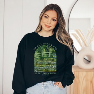 K&ouml;nnte beinhalten: Schwarzes Sweatshirt mit einem gr&uuml;nen und wei&szlig;en Bild einer Br&uuml;cke &uuml;ber einem Teich mit Seerosen. Der Text "He will make a way in the wilderness Isaiah 43:19" ist auf dem Sweatshirt aufgedruckt.