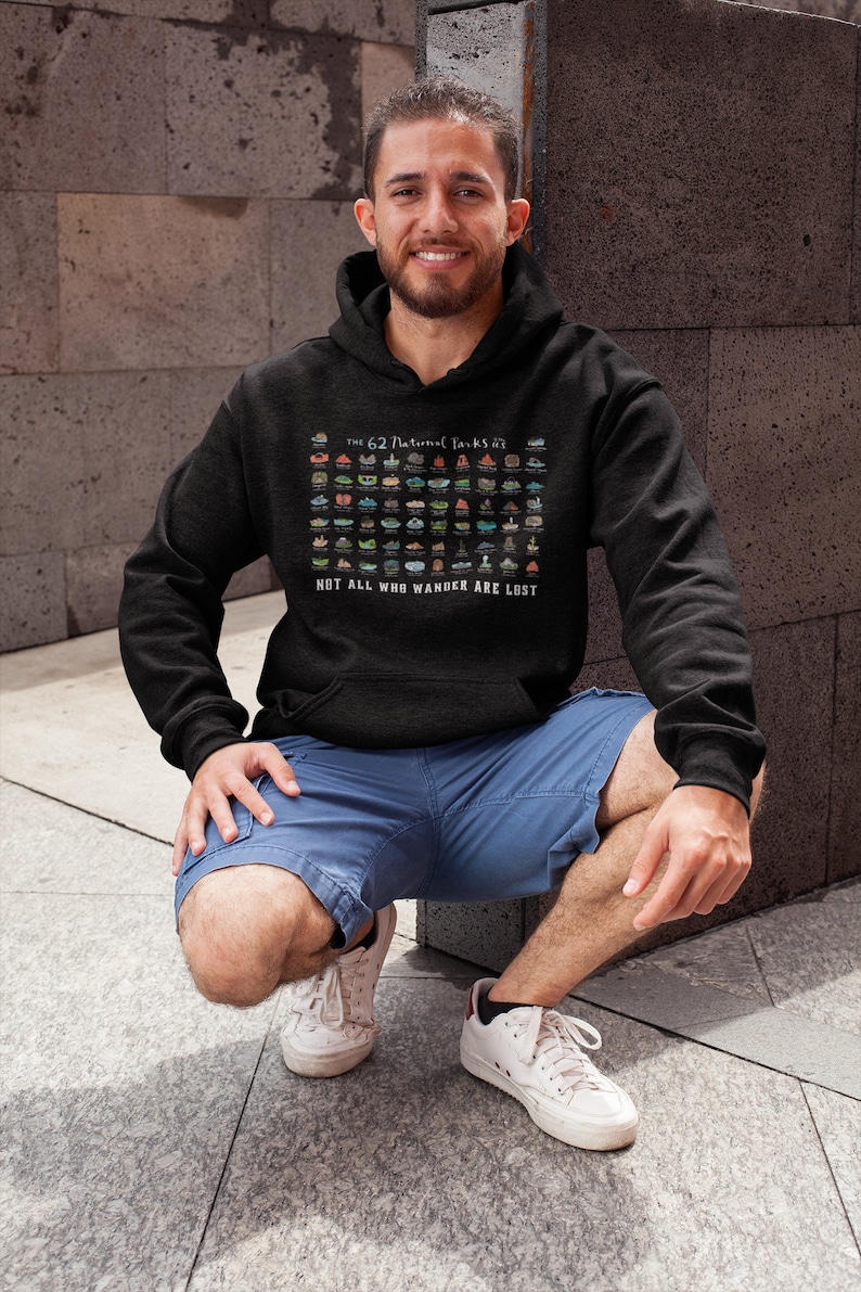 May include: A person wearing a black hoodie with a graphic of the 62 National Parks of the United States. The graphic is in color and includes the text "The 62 National Parks of the US" and "Not all who wander are lost".