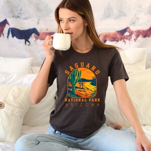 Pu&ograve; includere: Una maglietta marrone con una grafica in stile vintage di un paesaggio desertico con cactus saguaro. Il testo "Saguaro National Park Arizona" &egrave; stampato sotto la grafica.