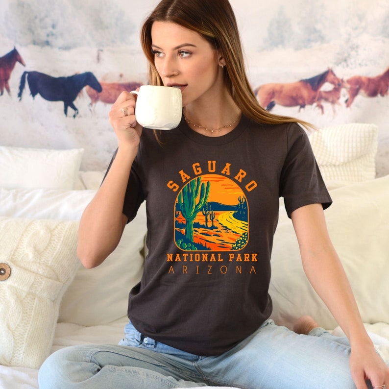 Pu&ograve; includere: Una maglietta marrone con una grafica in stile vintage di un paesaggio desertico con cactus saguaro. Il testo "Saguaro National Park Arizona" &egrave; stampato sotto la grafica.