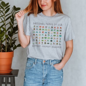 May include: A gray t-shirt with a graphic of all the National Parks of the USA. The graphic includes the names of each park and a small illustration of a landmark from each park. The text at the bottom of the graphic reads "SUPPORT - PROTECT - DISCOVER".