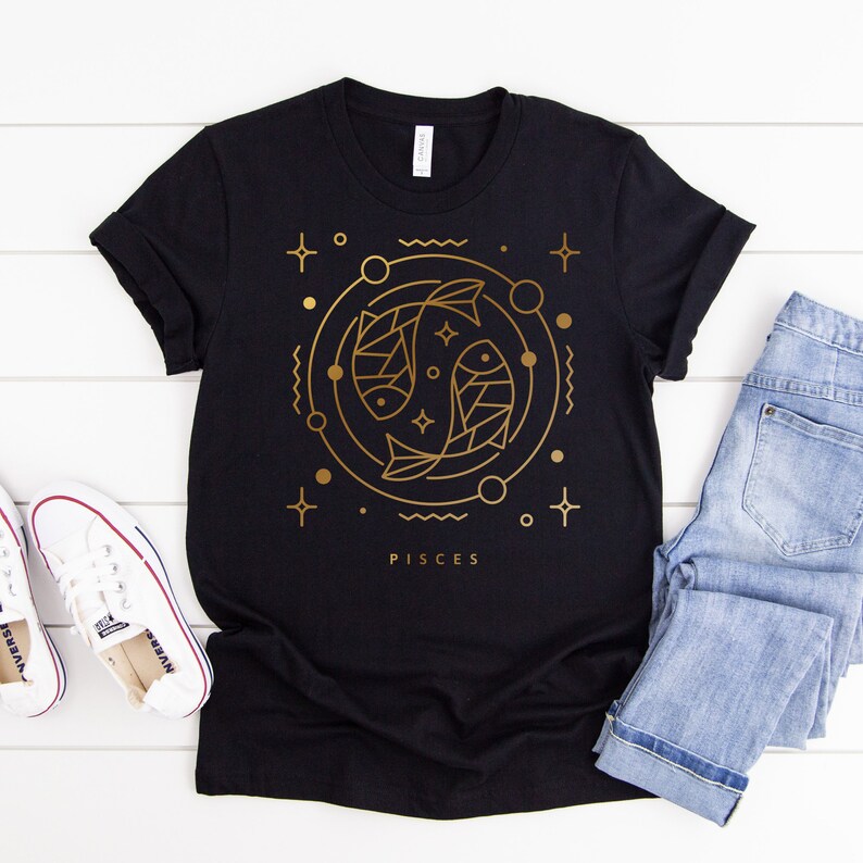 May include: Black t-shirt with a gold outline of two fish, a circle, and stars. The text "Pisces" is below the design.