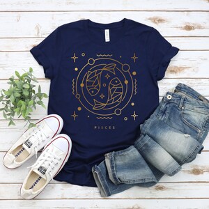 May include: A navy blue t-shirt with a gold outline of two fish and the word "Pisces" printed on the front. The shirt is laid flat on a wooden surface with a pair of white sneakers and blue jeans.