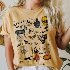 May include: A yellow t-shirt with a black and white graphic print of a skull, a goat, a bat, a moth, a spider web, a witch, a pumpkin, and the text "Something wicked this way comes."