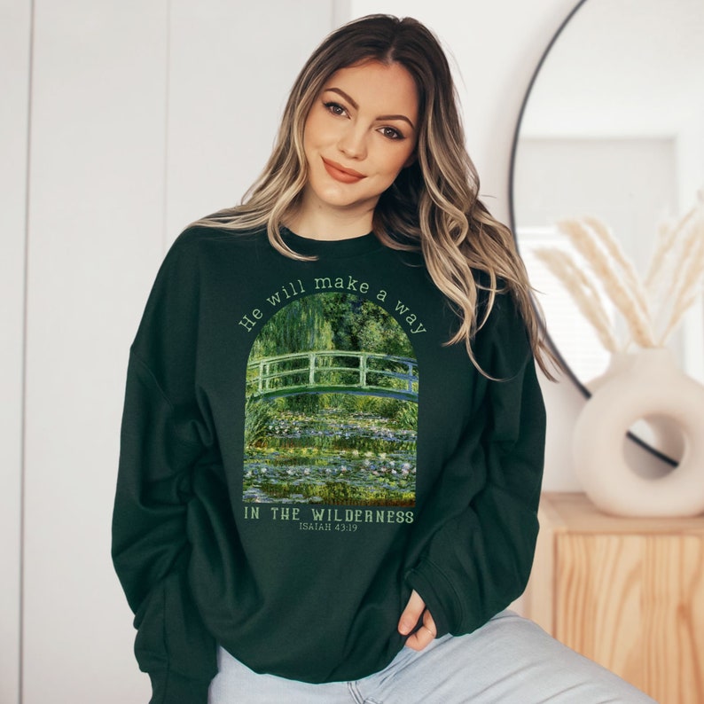 K&ouml;nnte beinhalten: Gr&uuml;nes Sweatshirt mit einem Gem&auml;lde einer Br&uuml;cke &uuml;ber einem Teich mit Seerosen. Der Text "He will make a way in the wilderness Isaiah 43:19" ist auf dem Sweatshirt aufgedruckt.