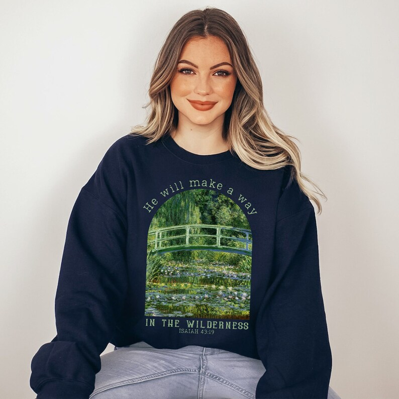 K&ouml;nnte beinhalten: Navyblauer Sweatshirt mit einem gr&uuml;nen und wei&szlig;en Gem&auml;lde einer Br&uuml;cke &uuml;ber einem Teich mit Seerosen. Der Text "He will make a way in the wilderness Isaiah 43:19" ist auf dem Sweatshirt aufgedruckt.