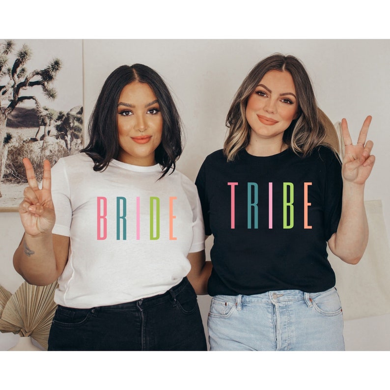 以下が含まれることがあります： 2人ともお揃いのTシャツを着ています。1枚は白地に虹色の「BRIDE」と書いてあります。もう1枚は黒地に虹色の「TRIBE」と書いてあります。