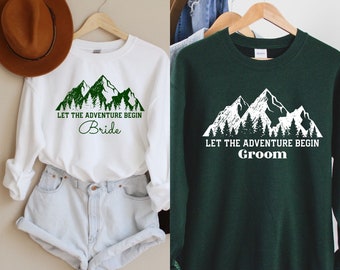 May include: Two sweatshirts with a mountain range graphic and the text "Let the adventure begin" with "Bride" on one and "Groom" on the other.