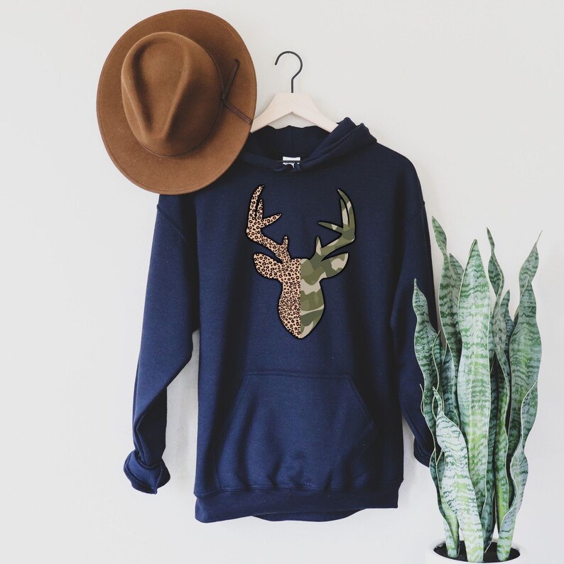 May include: A navy blue hooded sweatshirt with a deer silhouette design. The deer is split in half, with one side featuring a leopard print and the other side featuring a camouflage print.