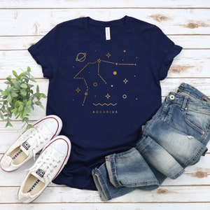 May include: A navy blue t-shirt with a gold outline of the Aquarius constellation and the word "Aquarius" printed below. The shirt is folded and laid on a white wooden surface with a pair of white sneakers and blue jeans.