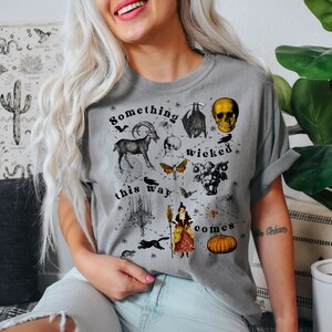 May include: A gray t-shirt with a black and white graphic print featuring a skull, a goat, a witch, a pumpkin, and the text "Something wicked this way comes".