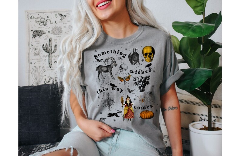 May include: A gray t-shirt with a black and white graphic print featuring a skull, a goat, a witch, a pumpkin, and the text "Something wicked this way comes".