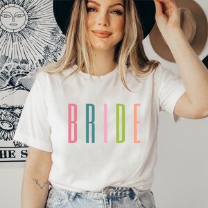 以下が含まれることがあります： 白いTシャツに「BRIDE」という単語がカラフルな文字でプリントされています。文字はピンク、ターコイズ、薄いピンク、グリーン、オレンジです。