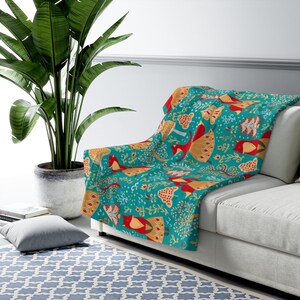 May include: A teal blue throw blanket with a repeating pattern of stylized birds, trees, and flowers in red, yellow, and orange. The blanket is draped over a white couch.