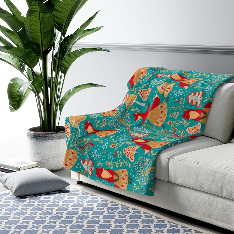 May include: A teal blue throw blanket with a repeating pattern of stylized birds, trees, and flowers in red, yellow, and orange. The blanket is draped over a white couch.