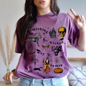 May include: Purple t-shirt with a black and white graphic print of a skull, bat, goat, moth, spider, and other spooky imagery. The text on the shirt reads "Something wicked this way comes."