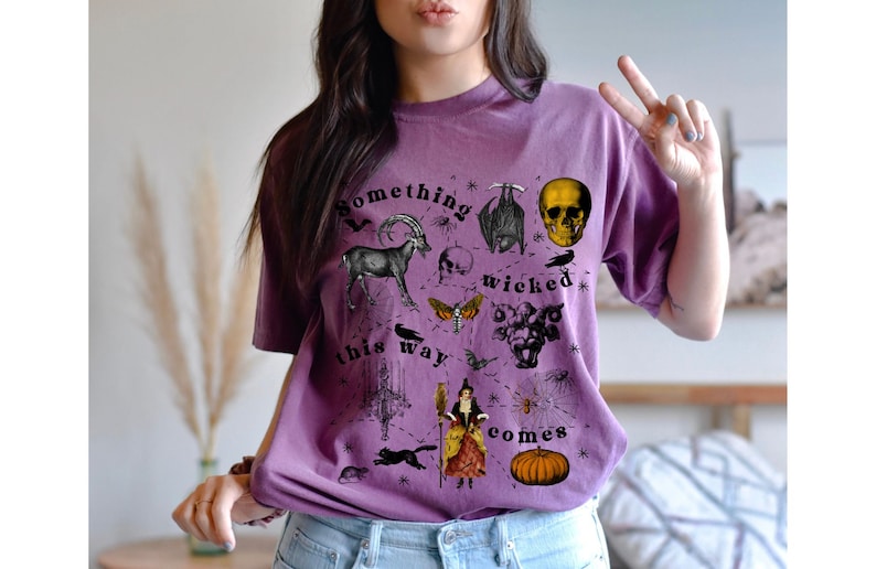 May include: Purple t-shirt with a black and white graphic print of a skull, bat, goat, moth, spider, and other spooky imagery. The text on the shirt reads "Something wicked this way comes."