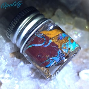 May include: A small glass vial containing a piece of iridescent opal with a red, brown, and gold matrix. The vial has a silver lid.