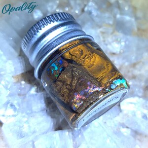 Könnte beinhalten: Ein kleines Glasgefäß mit einem silbernen Deckel, das ein Stück irisierenden Opals mit braunen und goldenen Markierungen enthält. Das Gefäß ist mit "SIN" beschriftet.