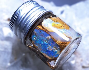 May include: A small glass vial with a silver lid containing a piece of blue and gold opal. The opal is iridescent and has a variety of colors.