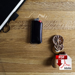 May include: A black leather lighter case with yellow stitching. The case is on a wooden surface.