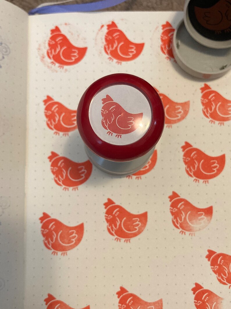 May include: A red rubber stamp with a white outline and a red chicken design. The stamp is on a white surface with multiple red chicken stamps.