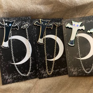 Puede incluir: Tres pines de esmalte con una criatura parecida a un murciélago con una luna creciente y una cadena. Los pines están en tarjetas negras con el texto "Gracias por su apoyo".