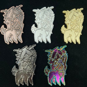 May include: Five enamel pins featuring a cartoon deer with a heart on its chest. The pins are in different colors: copper, silver, gold, black, and iridescent.