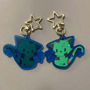 May include: Two blue and green cat keychains with gold star charms. The cats are cartoonish and have stitched-up wounds.