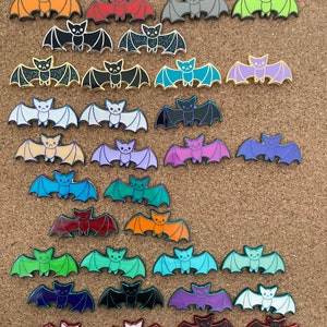 May include: A collection of 36 enamel pins featuring a cartoon bat in various colors, including orange, red, gray, green, blue, purple, white, and black. The bats have glitter accents and are arranged in rows on a corkboard.