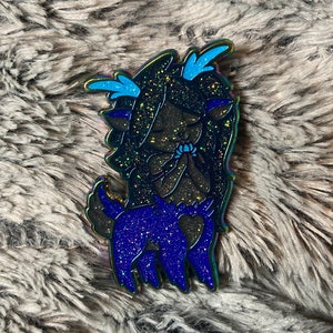 May include: A glittery blue and black enamel pin featuring a cartoon character with long black hair, blue eyes, and antlers. The character is wearing a blue dress with a heart-shaped design.