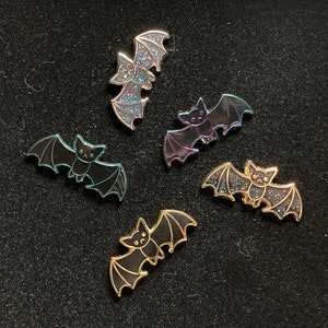 Può includere: Cinque spille a forma di pipistrello con glitter iridescenti. I pipistrelli sono in varie pose con le ali spiegate e ripiegate. Le spille sono di colore blu scuro, viola e oro.