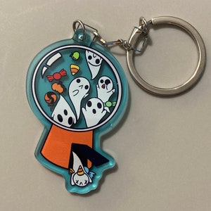 May include: A blue and orange acrylic keychain featuring a ghost-shaped gumball machine filled with candy and ghosts. A small ghost is hanging upside down from the bottom of the gumball machine.