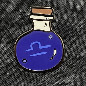 May include: A blue enamel pin shaped like a potion bottle with a cork stopper. The bottle has a light blue Libra symbol inside.