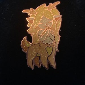 May include: A gold glitter enamel pin of a cartoon deer with a heart on its chest. The deer has long, flowing hair and is holding its hands together in a prayer position.