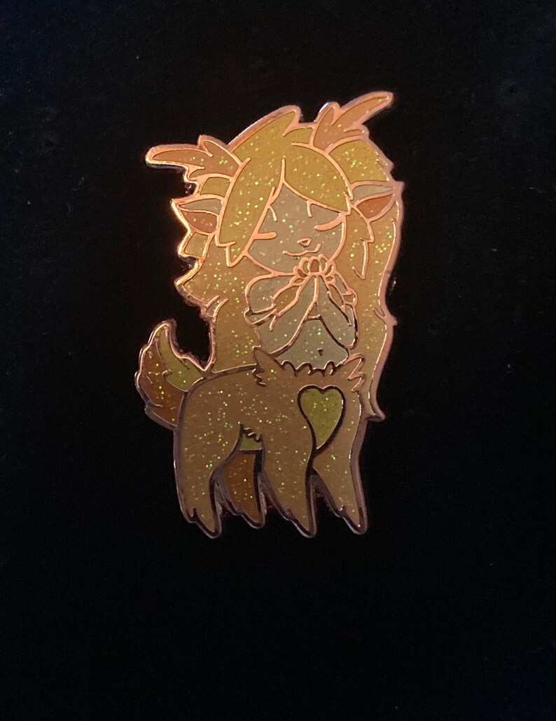 May include: A gold glitter enamel pin of a cartoon deer with a heart on its chest. The deer has long, flowing hair and is holding its hands together in a prayer position.