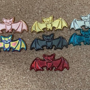May include: Seven different colored enamel pins shaped like bats. The bats are all the same size and have a shiny finish. The colors are pink, gold, silver, teal, black, red, and yellow.