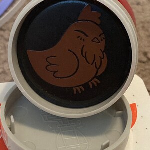 May include: A round, gray and red plastic stamp with a black and brown chicken design. The chicken is facing left and has a red comb.