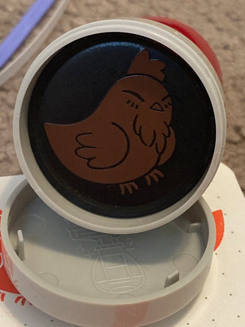 May include: A round, gray and red plastic stamp with a black and brown chicken design. The chicken is facing left and has a red comb.