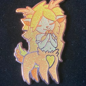 May include: A gold glitter enamel pin of a cartoon fawn with a heart on its back and long blonde hair. The fawn is holding a flower in its hands and has its eyes closed.