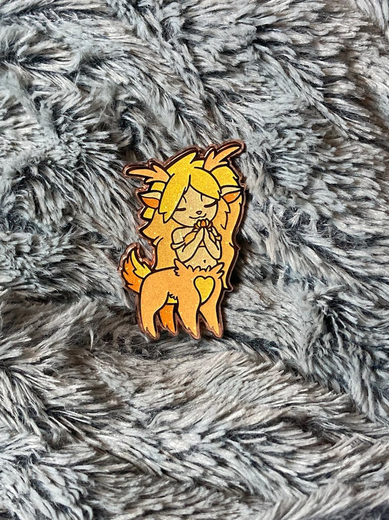 May include: A wooden pin of a cartoon fawn with a heart on its back. The fawn has yellow hair and is wearing a white dress. The pin is on a gray fuzzy surface.