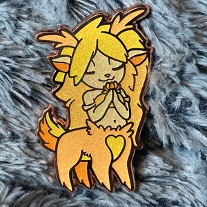 May include: A gold glitter enamel pin of a yellow and orange deer-like creature with a heart on its chest. The creature has a flower in its hands and is looking down.