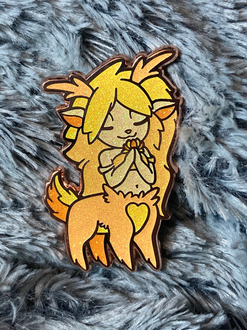 May include: A gold glitter enamel pin of a yellow and orange deer-like creature with a heart on its chest. The creature has a flower in its hands and is looking down.