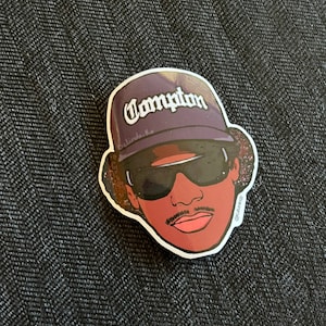 May include: A sticker of a cartoon illustration of a person wearing a purple baseball cap with the word "Compton" written on it. The person is wearing sunglasses and has a mustache and a pink mouth.
