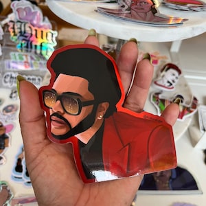 May include: A red and black sticker featuring a portrait of a man with a beard and sunglasses. The sticker has a glossy finish and the text "@Kulturalogie" is printed on the bottom right corner.