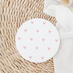 May include: A circular white coaster adorned with a scattering of small, pink hearts. It rests on a woven, light brown mat, with a white cloth and dried flowers in the background. The coaster is a decorative item, suitable for a home.