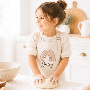 May include: A cream-coloured apron featuring a printed rainbow graphic in muted earth tones with the name Daisy written in black script below the rainbow. The apron has adjustable shoulder straps and a rectangular bib front design.