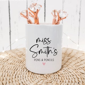 May include: White ceramic pencil holder with the text "miss Smith's PENS & PENCILS" and a pink heart. The holder has five rose gold pens with clear crystal tops.